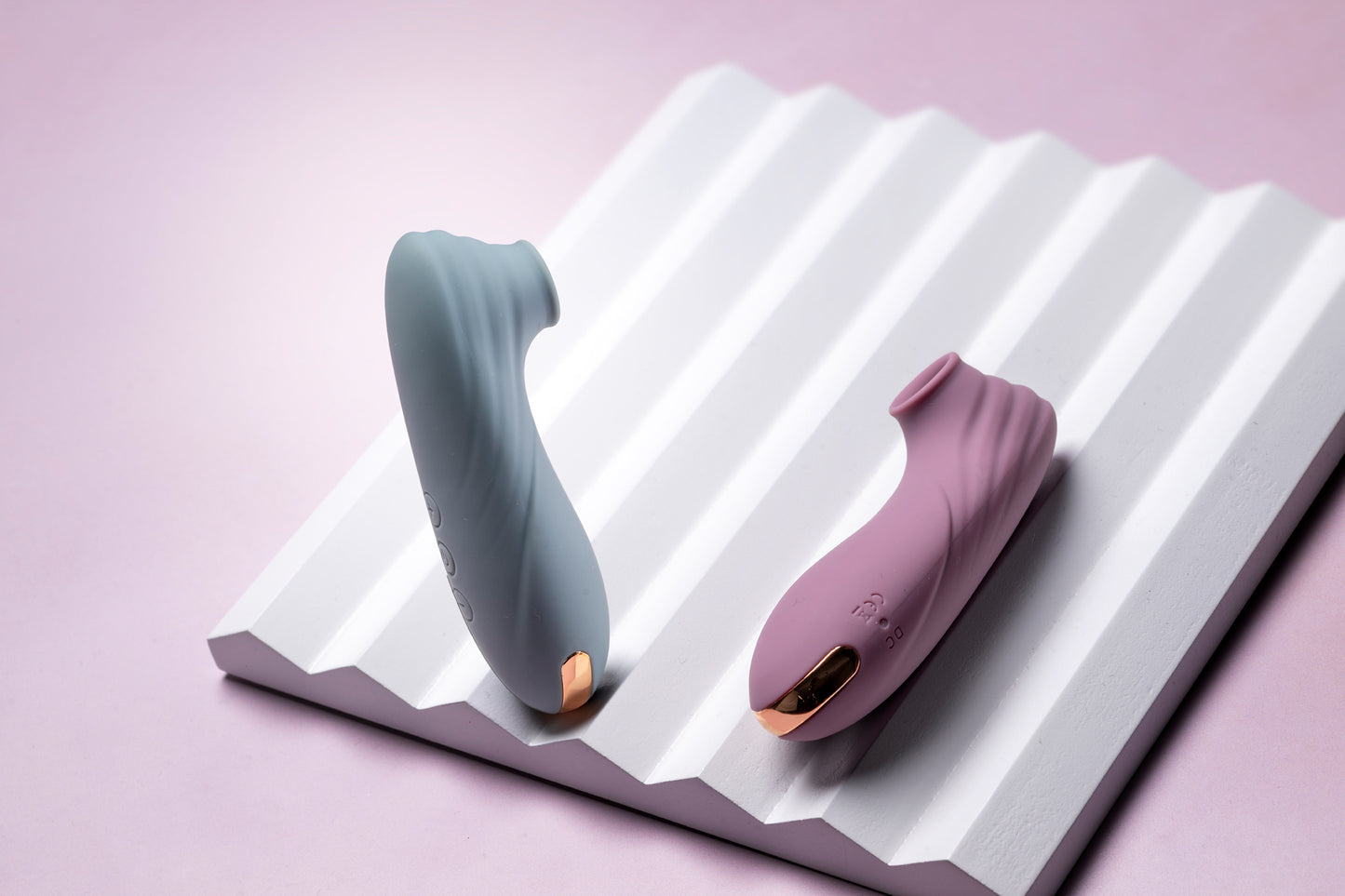 Pink clitoral suction vibrator laying down, teal one standing on white prop, pink background